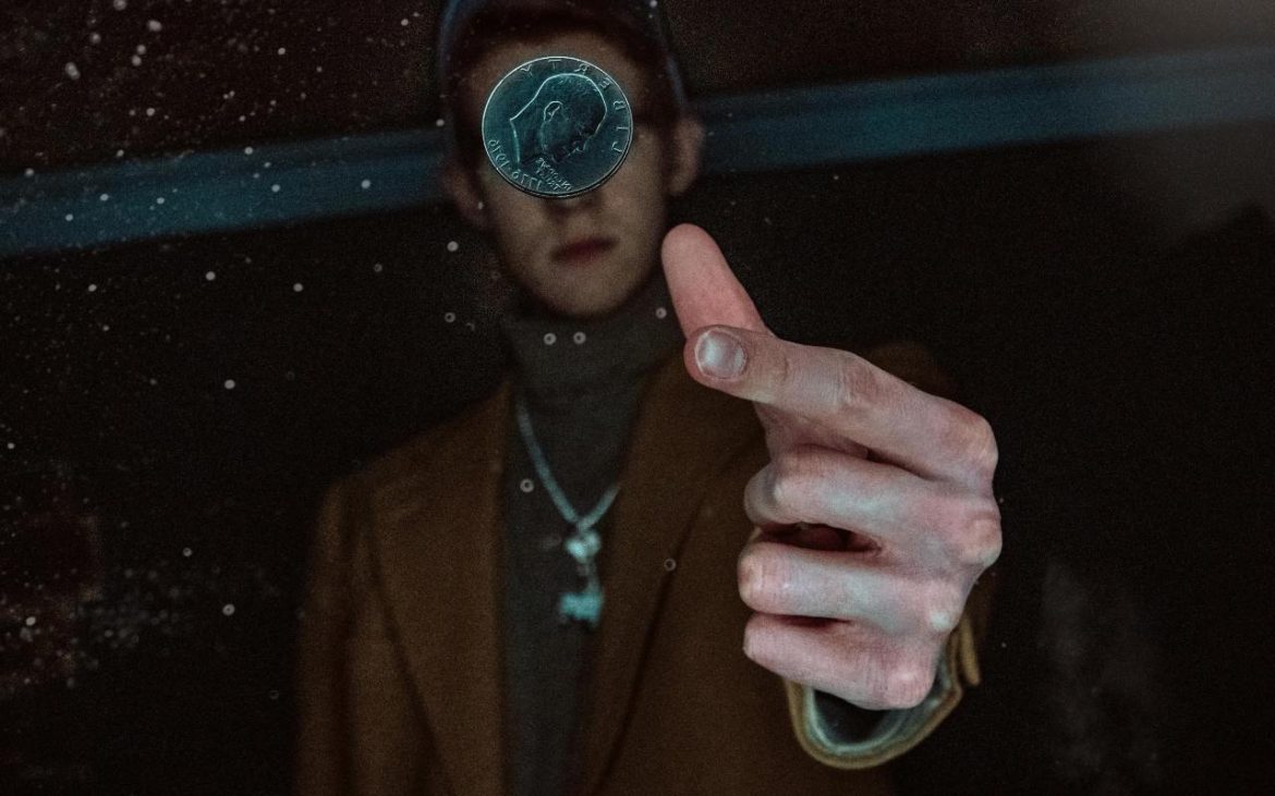 Čovek u braon sakou i kačketom baca novčić