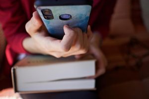 Ruke drže mobilni telefon iznad dve naslagane knjige